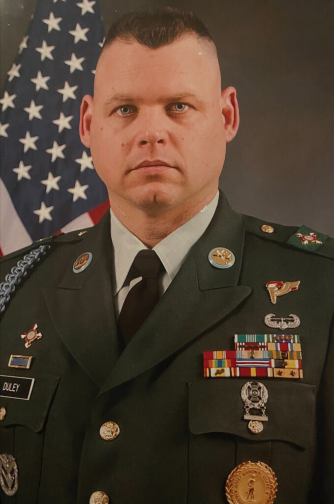 Formal military portrait of a soldier in Army green dress uniform with service ribbons and badges, American flag behind