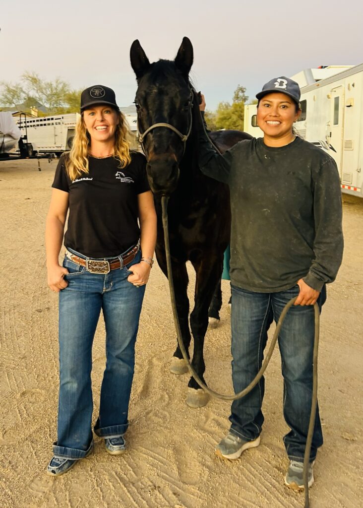 Two women in ball caps and jeans stand smiling beside a black horse in a halter and lead rope at a trailer parking area