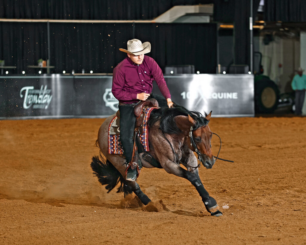 Rider in a maroon shirt and cowboy hat slides a bay roan horse to a stop in a dirt arena during a reining run