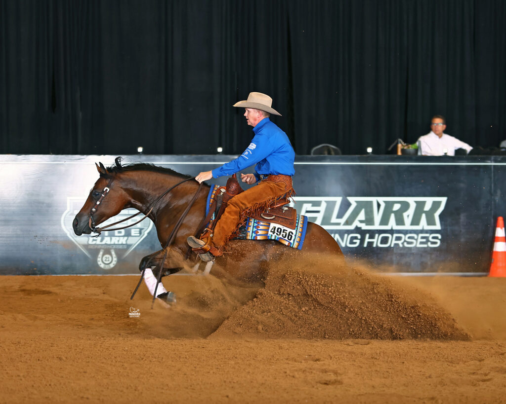 Reining horse wearing number 4956 slides to a stop in a spray of dirt as its rider in a blue shirt and chaps looks ahead