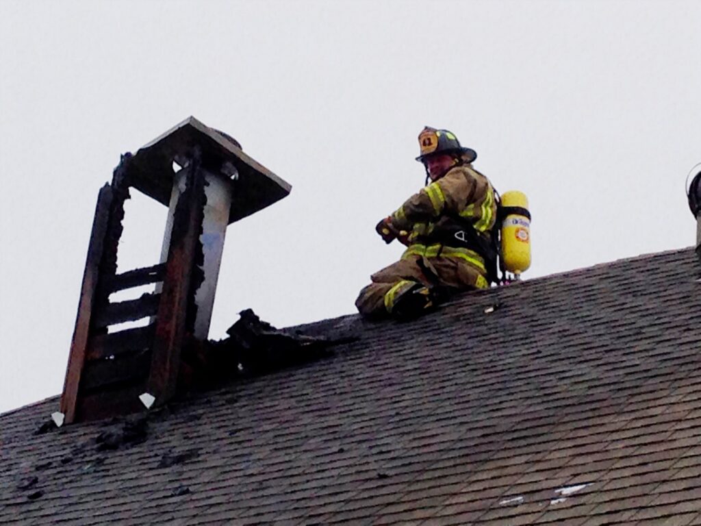 Firefighter in helmet and turnout gear kneels on a shingled roof beside a charred, burned-out cupola