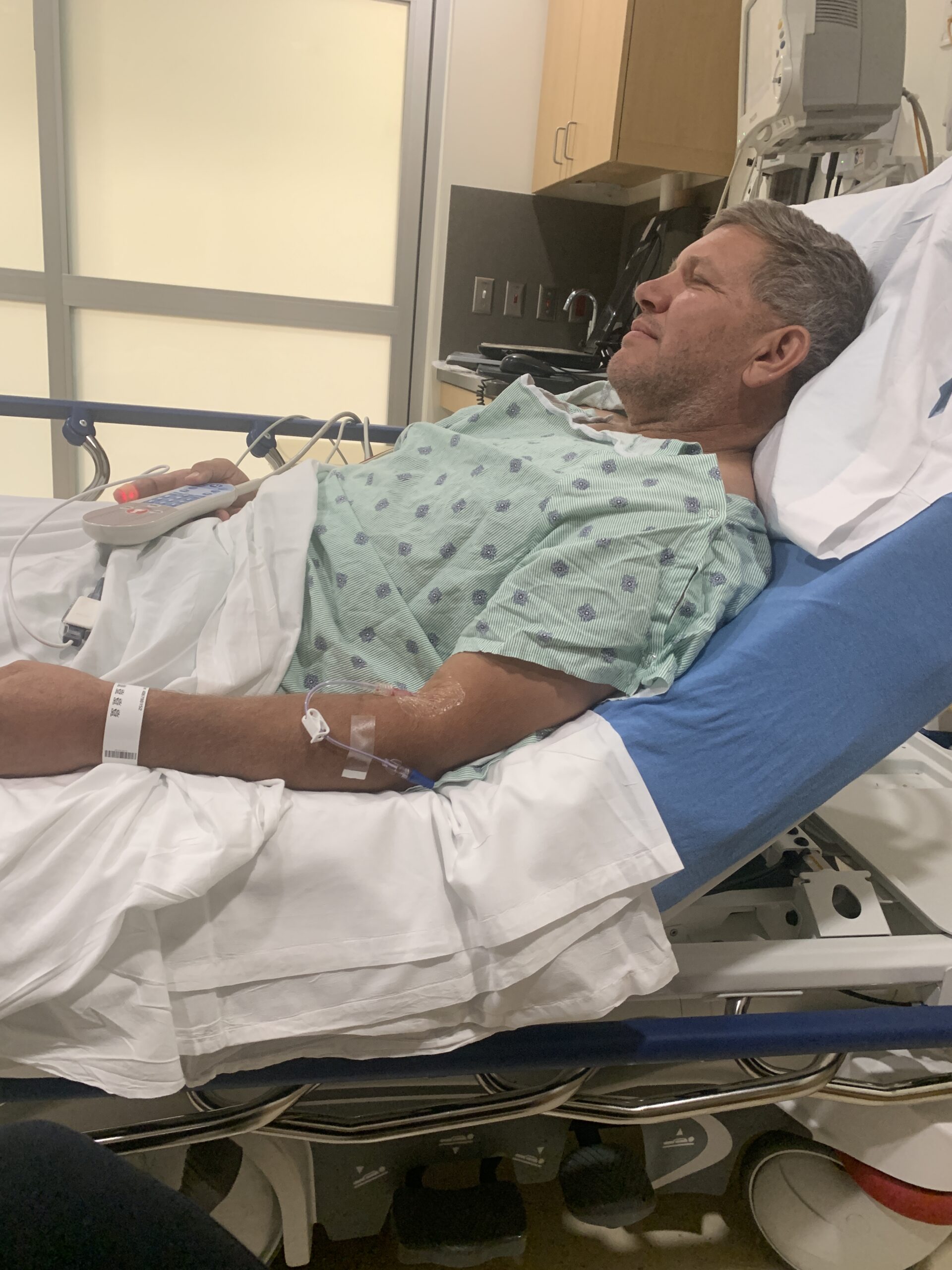 Gray-haired man in a hospital gown rests in a hospital bed with an IV in his arm and call remote in hand