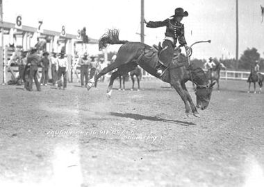 Cowboy riding a bucking horse during a rodeo event, with spectators and grandstands visible in the background