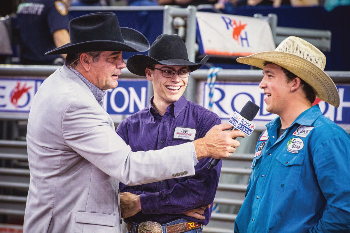 Announcer in light blazer and black cowboy hat interviews two rodeo competitors in western shirts and cowboy hats at arena sidelines