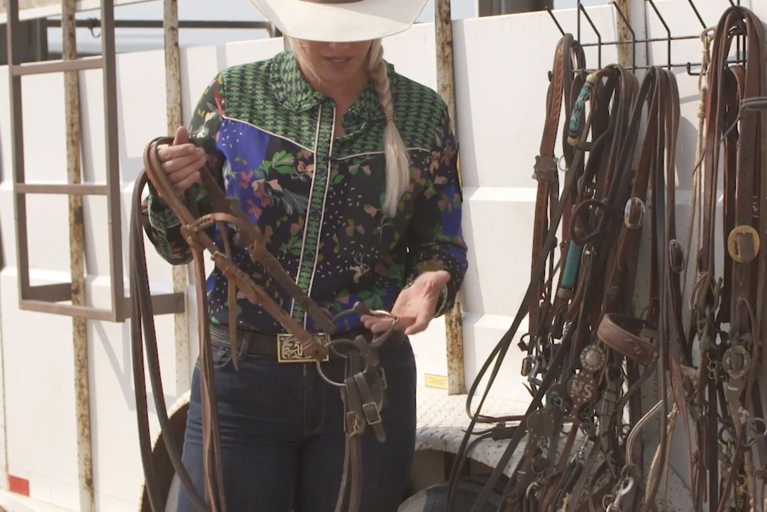 Woman in cowboy hat and floral western shirt holds a leather bridle with bit beside a trailer rack of hanging headstalls