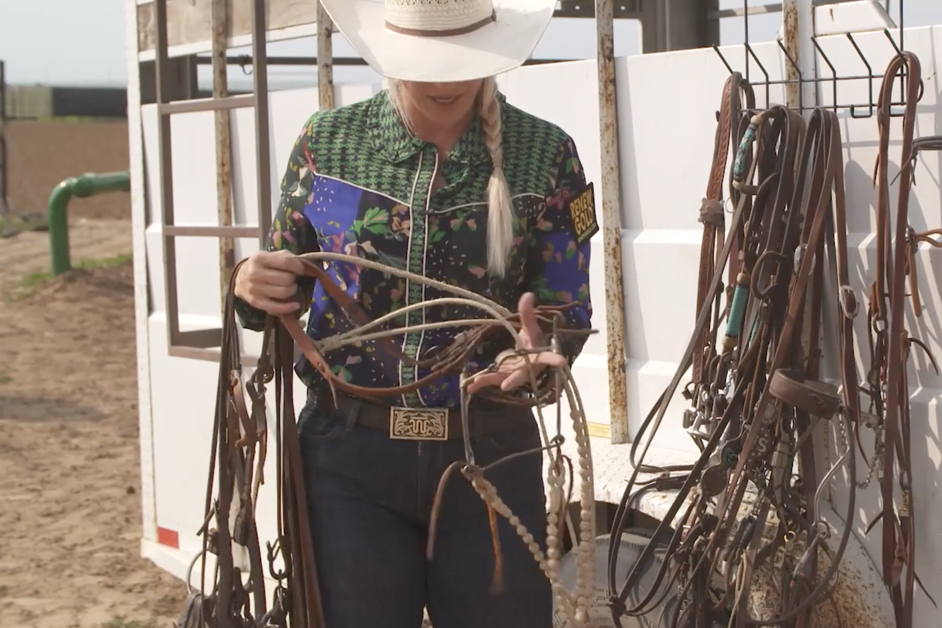 Cowgirl in a straw hat and floral western shirt sorts leather bridles and reins beside a trailer-mounted tack rack