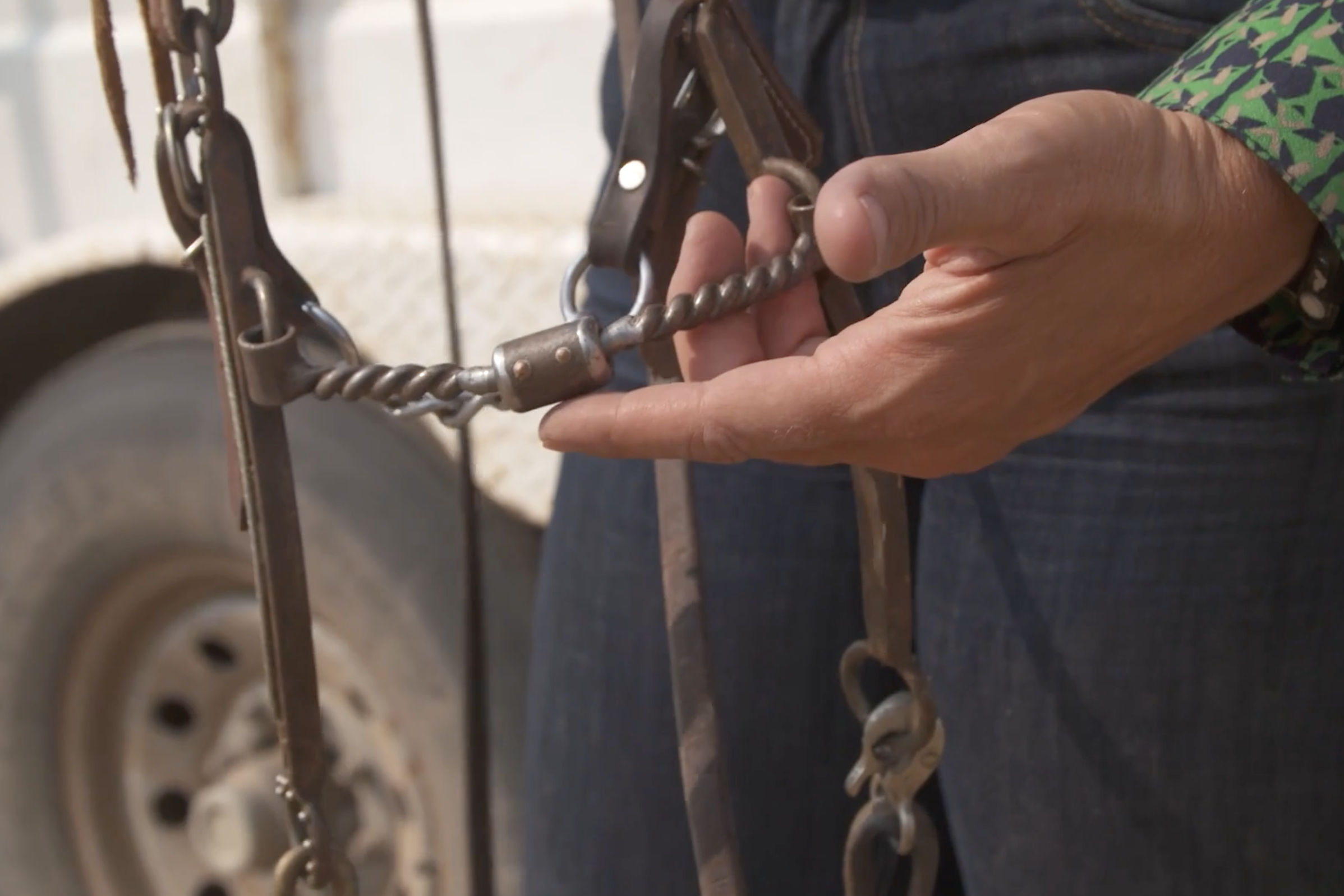 Close-up of a hand holding a twisted-wire snaffle bit attached to a leather headstall beside a trailer wheel