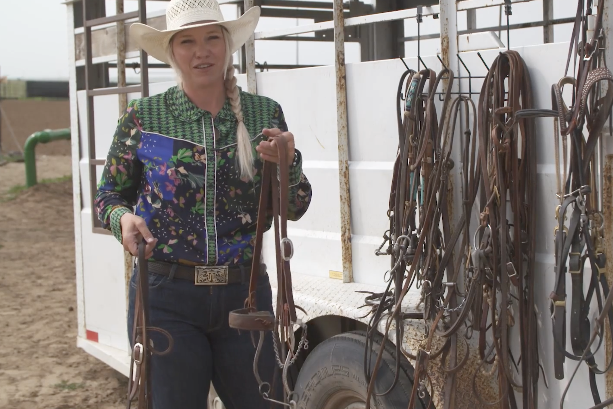 Woman in a straw cowboy hat and floral western shirt holds up bridles beside a trailer hung with bits and headstalls