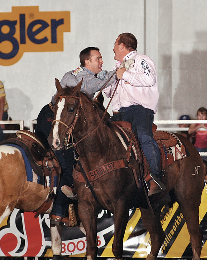 Two mounted cowboys grapple face-to-face on horseback in front of a Wrangler banner at an indoor arena