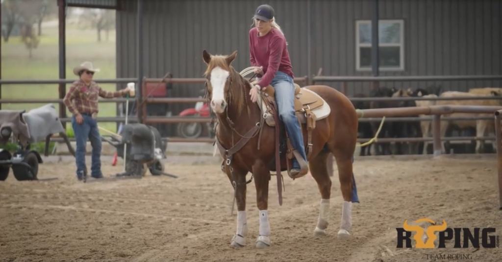 Rider in a red shirt and ball cap sits mounted on a sorrel horse in a sandy arena while a man in a cowboy hat watches from the fence