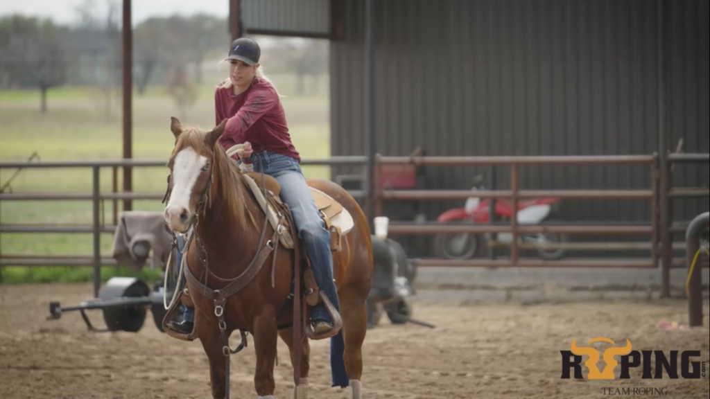 Young woman in a maroon shirt and ball cap rides a sorrel horse in a sandy arena beside a covered barn