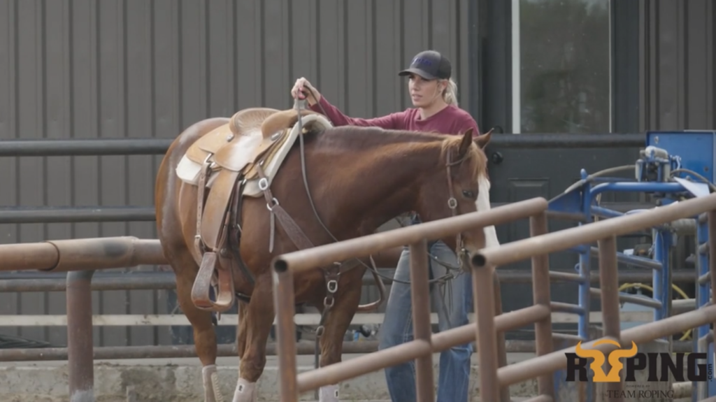 Woman in a ball cap adjusts the reins on a saddled sorrel horse in a pen outside a metal barn