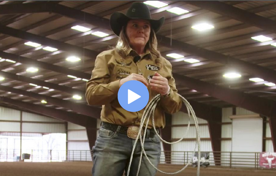Woman in a black cowboy hat and tan western shirt holding a coiled rope in an indoor arena, video play button overlaid