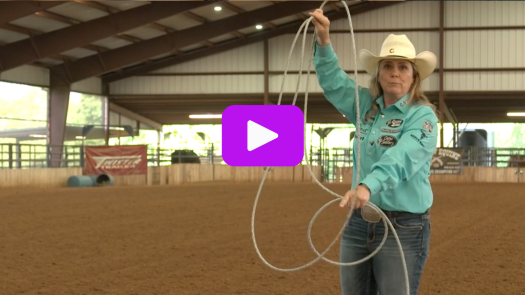 Breakaway roper in a teal shirt and straw cowboy hat swings her loop overhead in a covered dirt arena