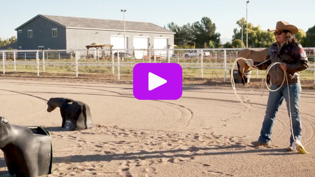 Cowgirl in a straw hat and sunglasses swings a loop toward a roping dummy in a sandy practice arena
