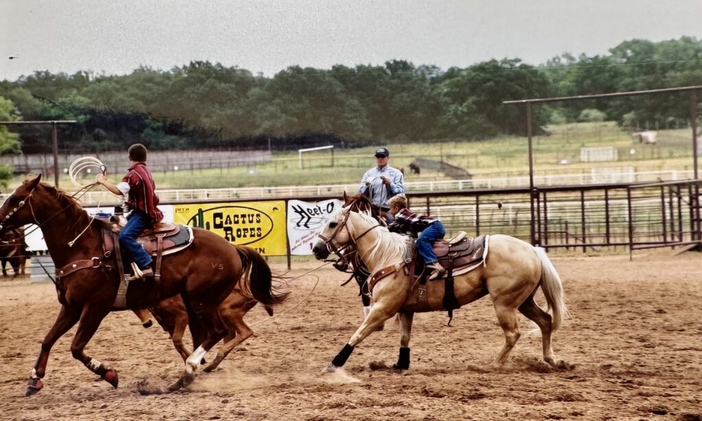 Two young team ropers gallop across a dirt arena past Cactus Ropes and Heel-O banners as a judge looks on