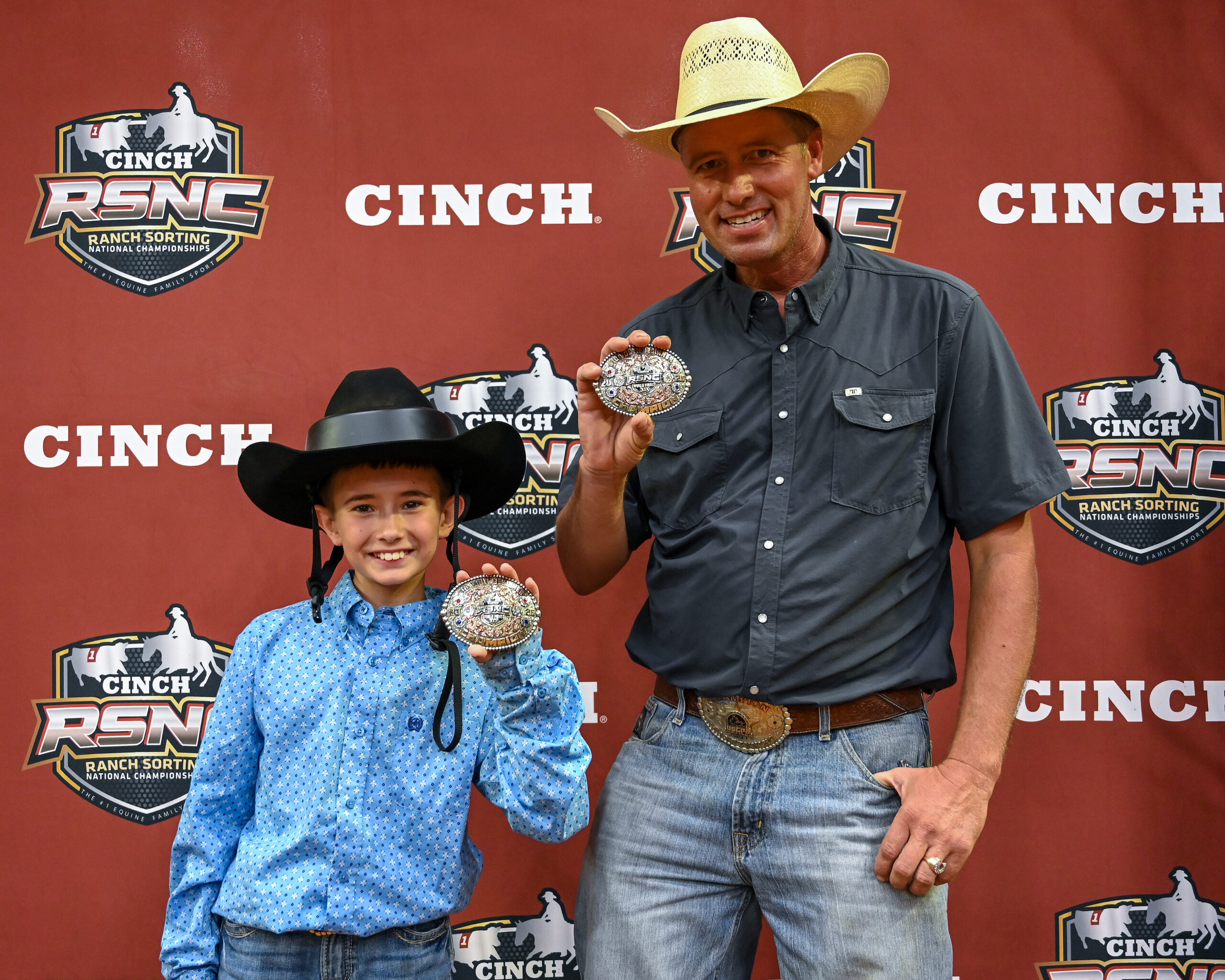 Man and child in Western attire hold championship buckles at Cinch RSNC Ranch Sorting National Championships event