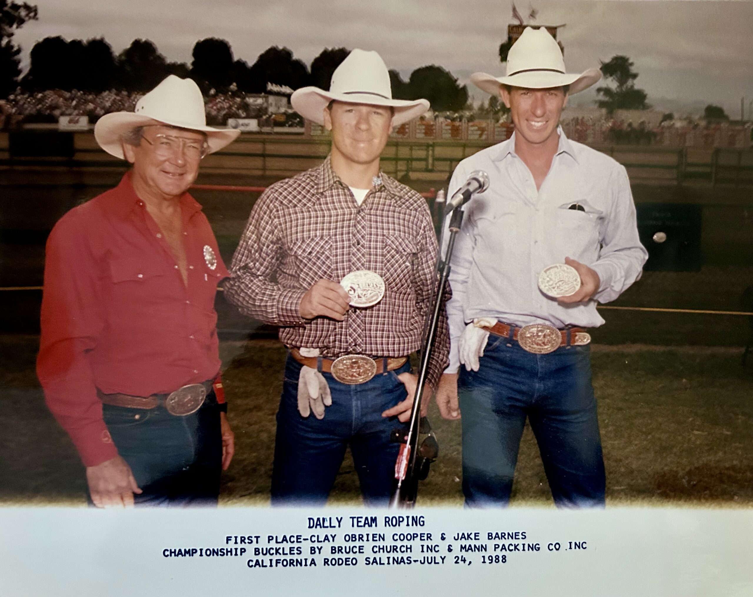 Three men in cowboy hats and Western wear display championship belt buckles at the 1988 California Rodeo Salinas team roping competition