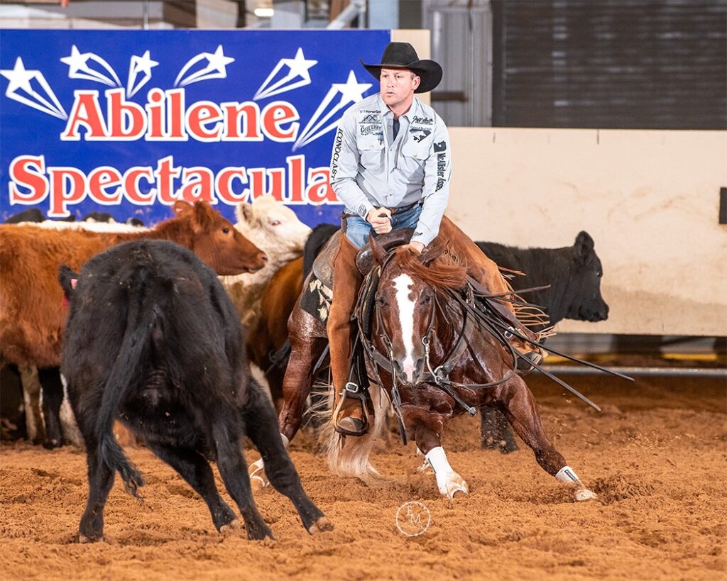 Cutting horse rider in black cowboy hat works a black cow in the arena dirt beneath an Abilene Spectacular banner