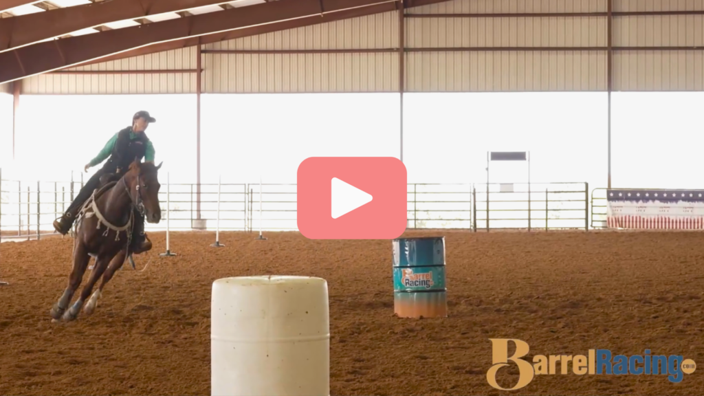 Rider in a green shirt and cap loping a sorrel horse through a covered arena past white and teal barrels