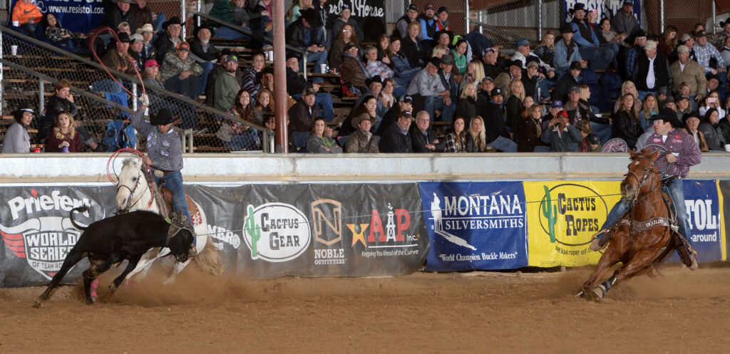 Team ropers work a steer down the arena fence past sponsor banners and packed grandstands, header swinging his loop