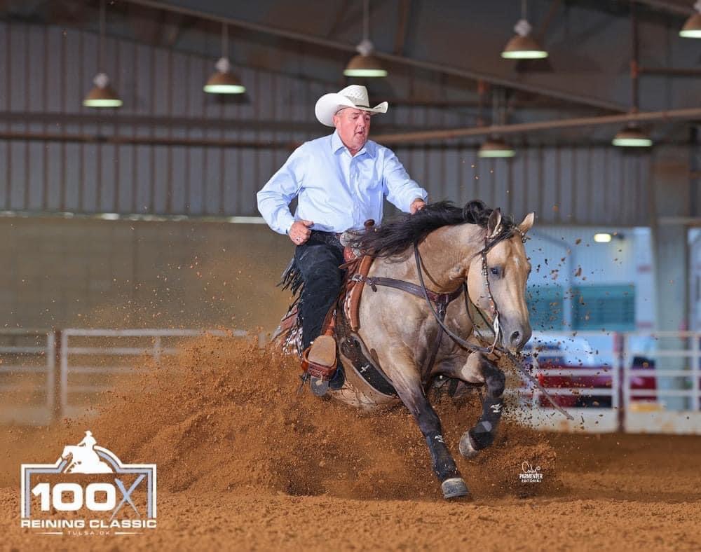 Reiner in a white cowboy hat and blue shirt slides his buckskin horse to a stop, kicking up dirt at the 100X Reining Classic