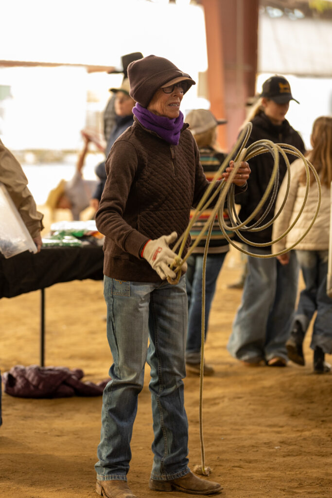 Woman in a knit cap, purple scarf and roping glove coils her lariat during a roping clinic in a dirt arena