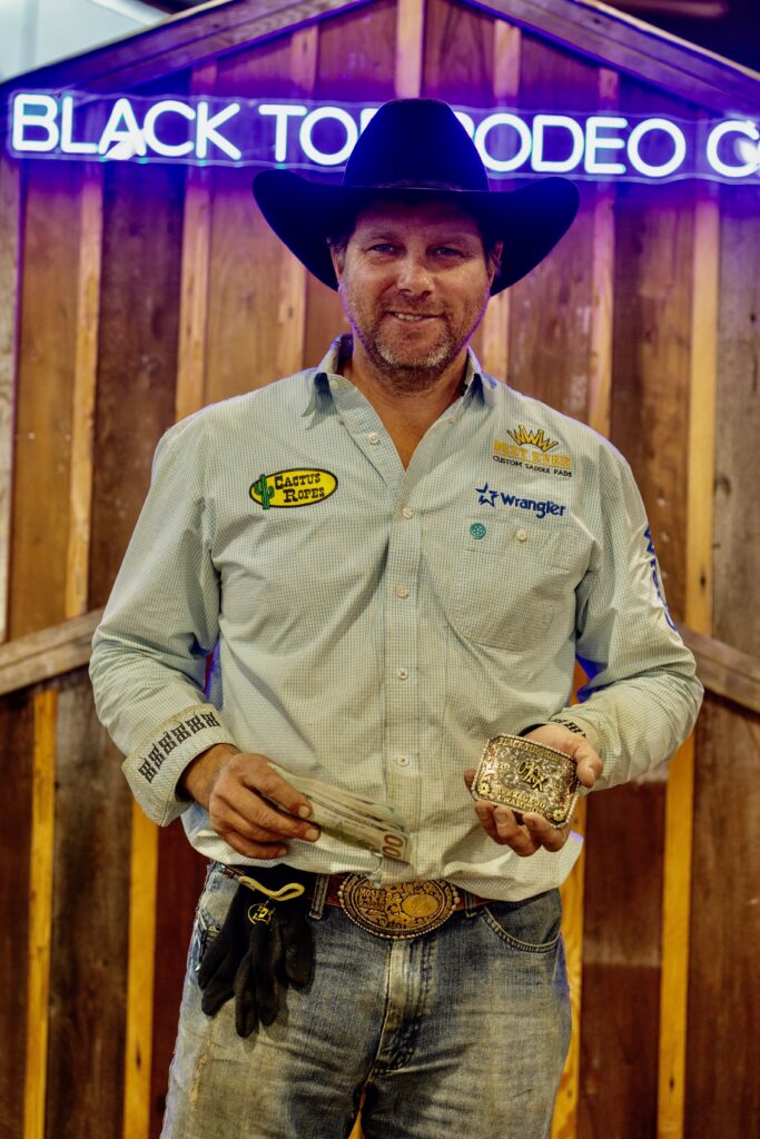Cowboy in a black hat holds a gold champion buckle and a fold of cash beneath a neon rodeo sign