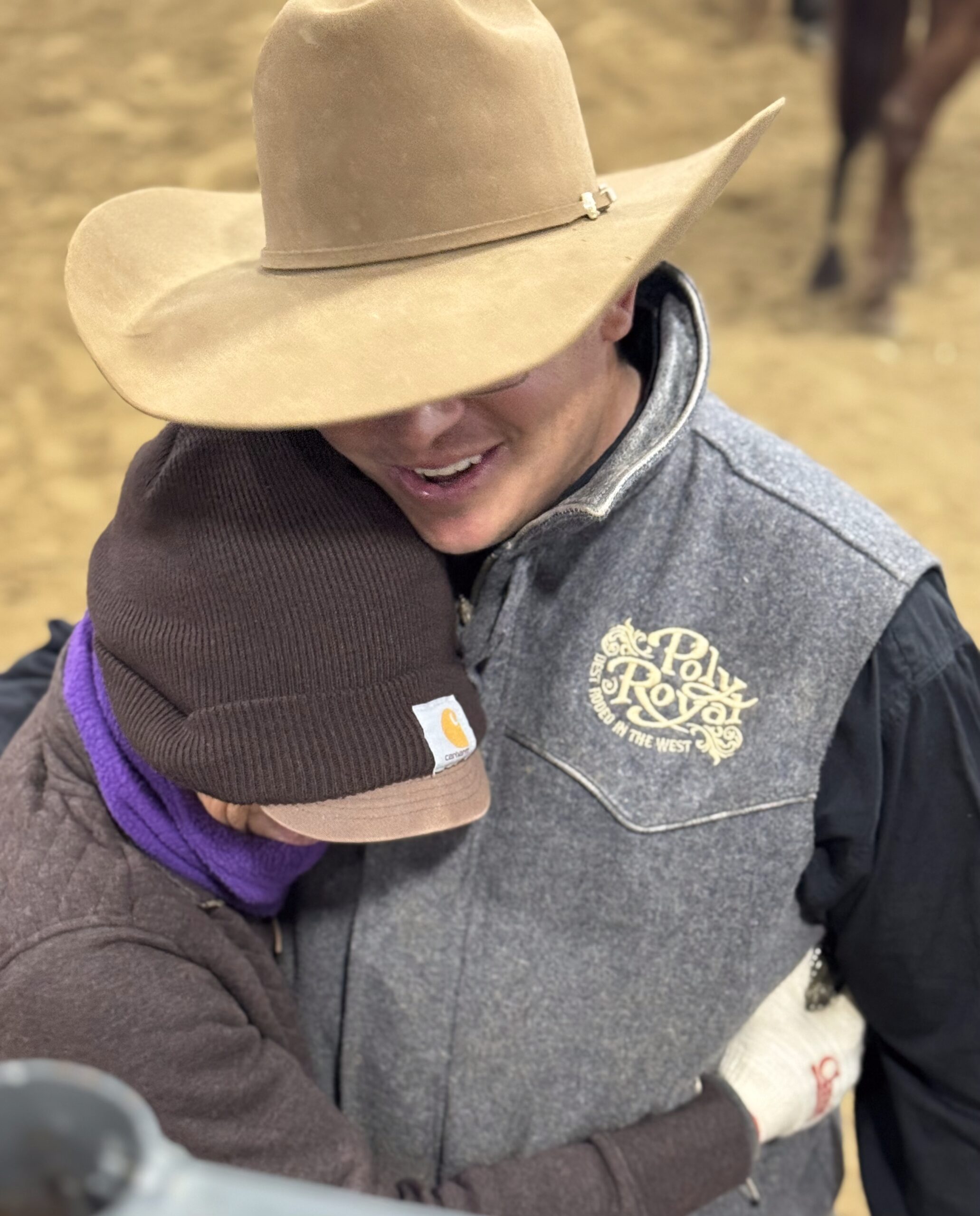 Smiling cowboy in a tan hat and gray "Poly Royal" rodeo vest hugs a person in a brown Carhartt beanie in an arena