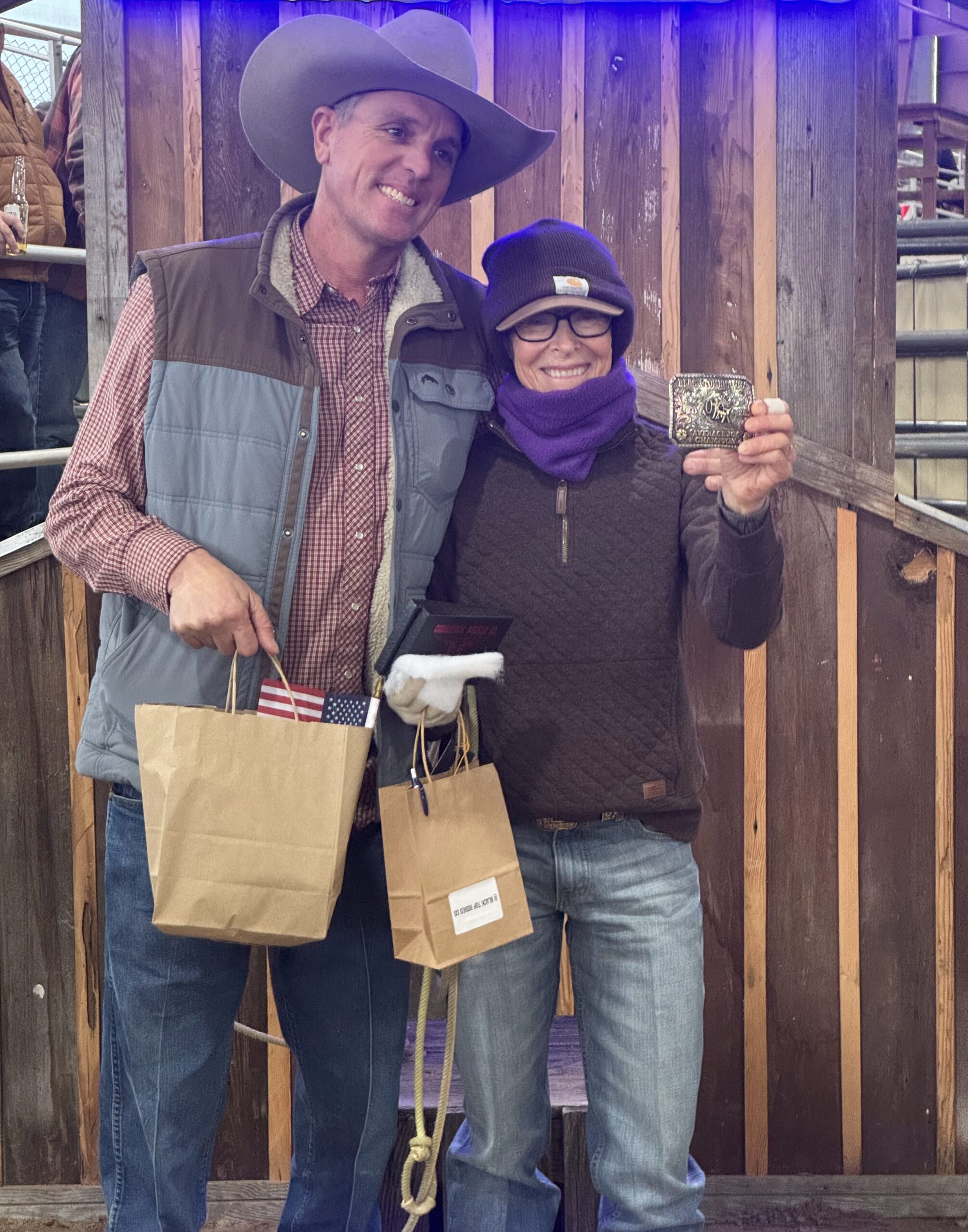 Smiling cowboy in a felt hat holding prize gift bags stands beside a woman in a beanie displaying a trophy buckle