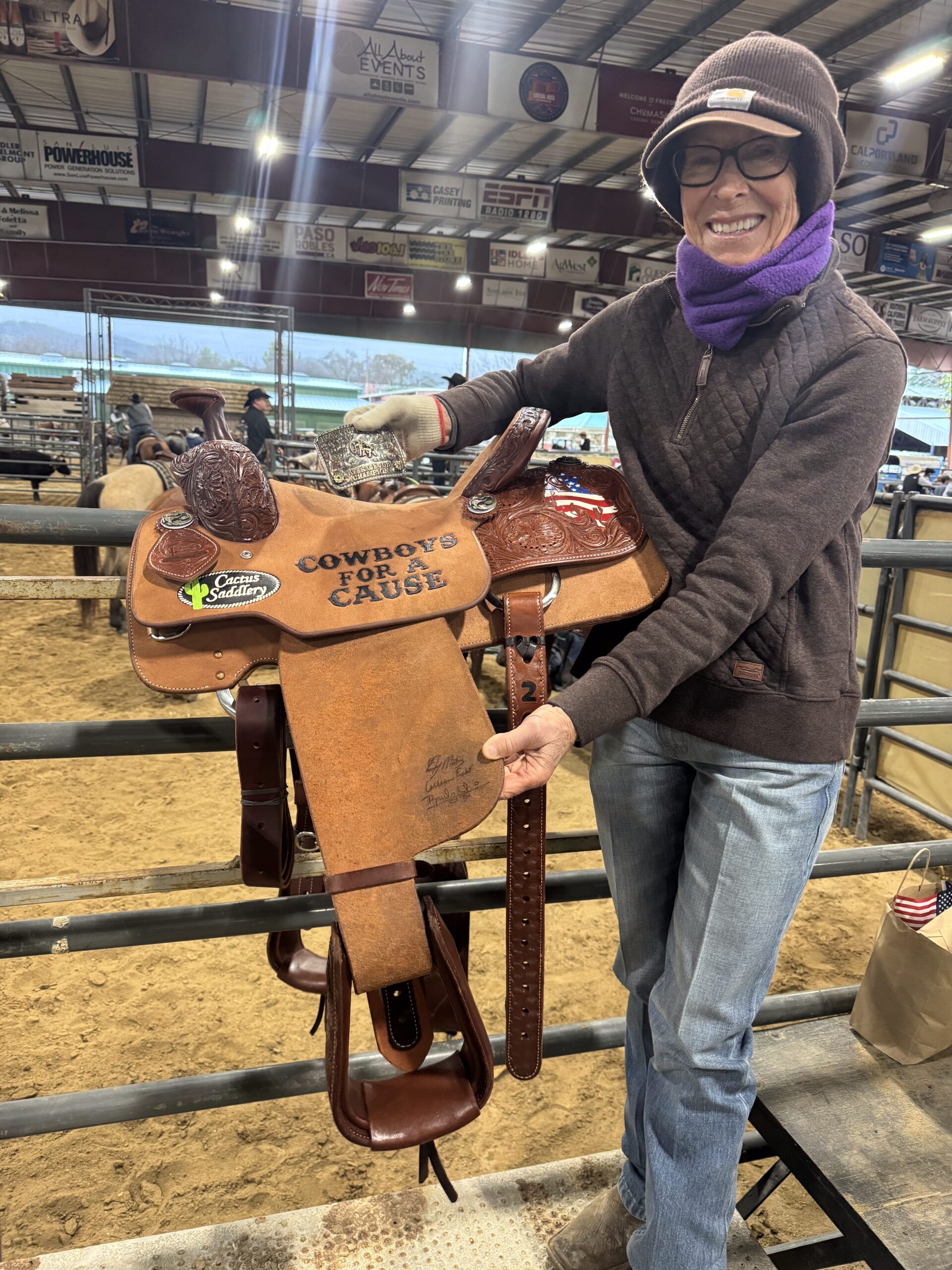 Smiling woman in beanie and purple neck gaiter holds a tooled "Cowboys for a Cause" Cactus Saddlery trophy saddle and belt buckle at an indoor arena