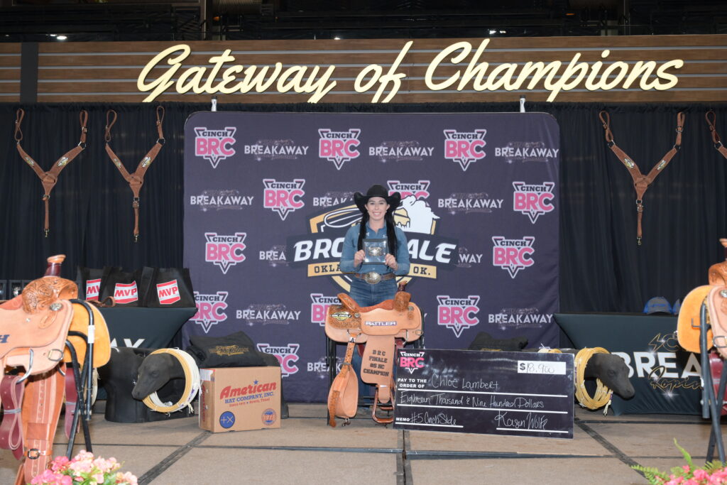 Breakaway roper in black cowboy hat holds a champion buckle onstage beside a trophy saddle and oversized check