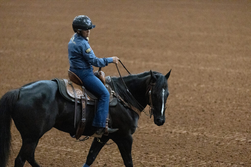 Rider in a blue shirt and helmet guides a black horse with a blaze at a walk across a dirt arena
