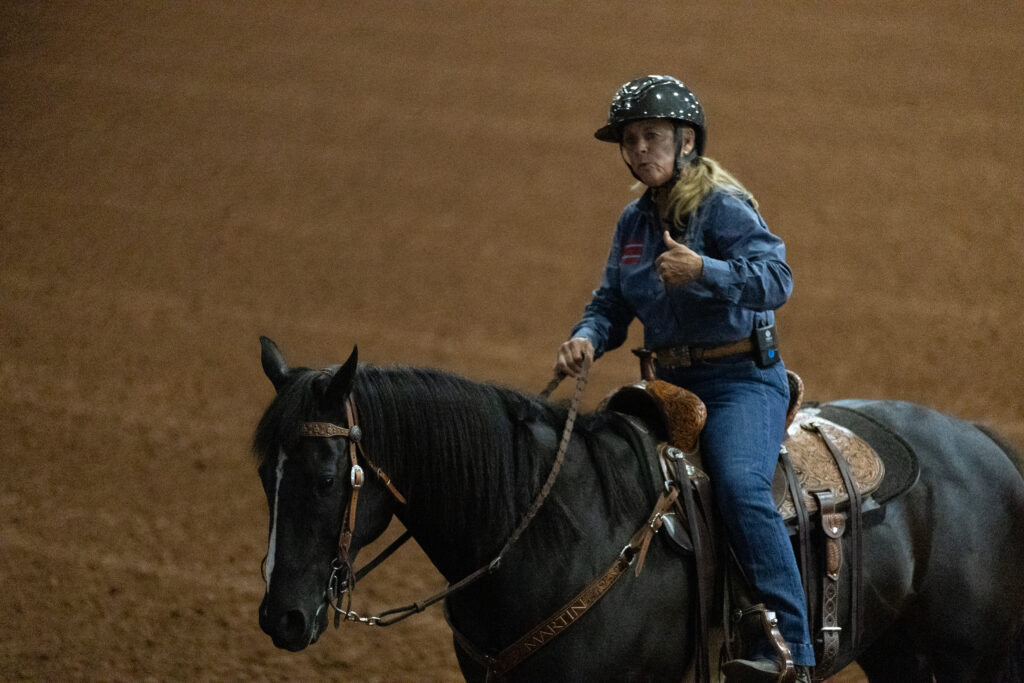 Rider in a blue shirt and helmet gives a thumbs-up while riding a black horse in a dirt arena