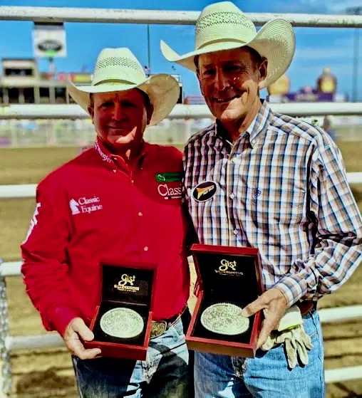 Two cowboys in western attire holding buckles and awards at a rodeo arena