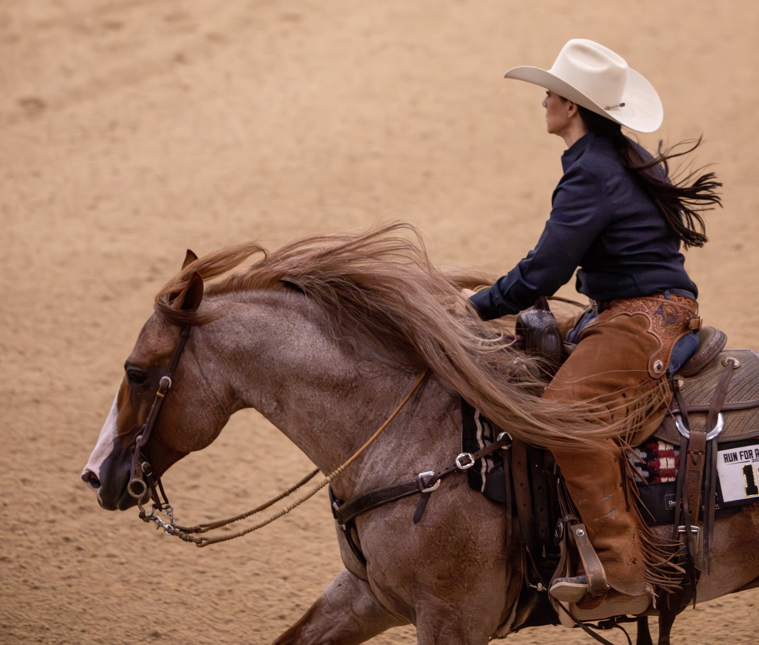 Rider in a white cowboy hat and leather chaps guides a roan horse with a flowing mane across the arena dirt