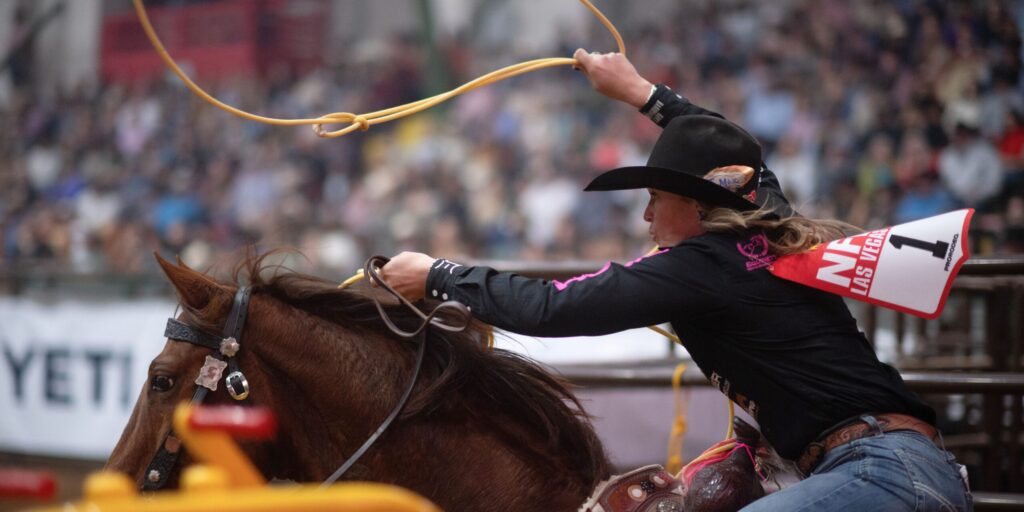 Breakaway roper in a black shirt swings her loop overhead aboard a sorrel horse at an indoor rodeo, back number 1 visible