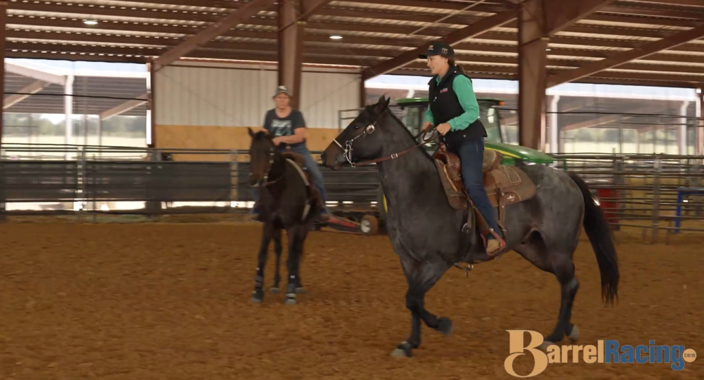Rider in a teal shirt and vest schools a blue roan horse in a covered arena while another rider watches from behind