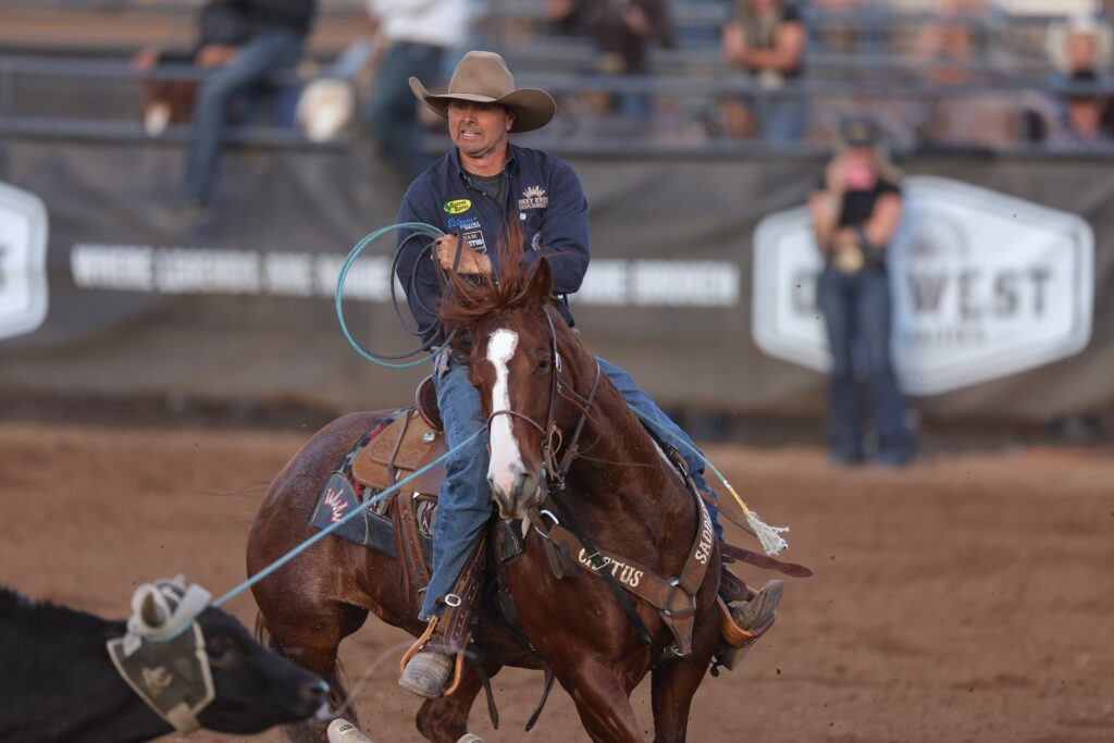 Cowgirl on horseback swinging a lasso during a rodeo event, with spectators visible in the grandstand behind her