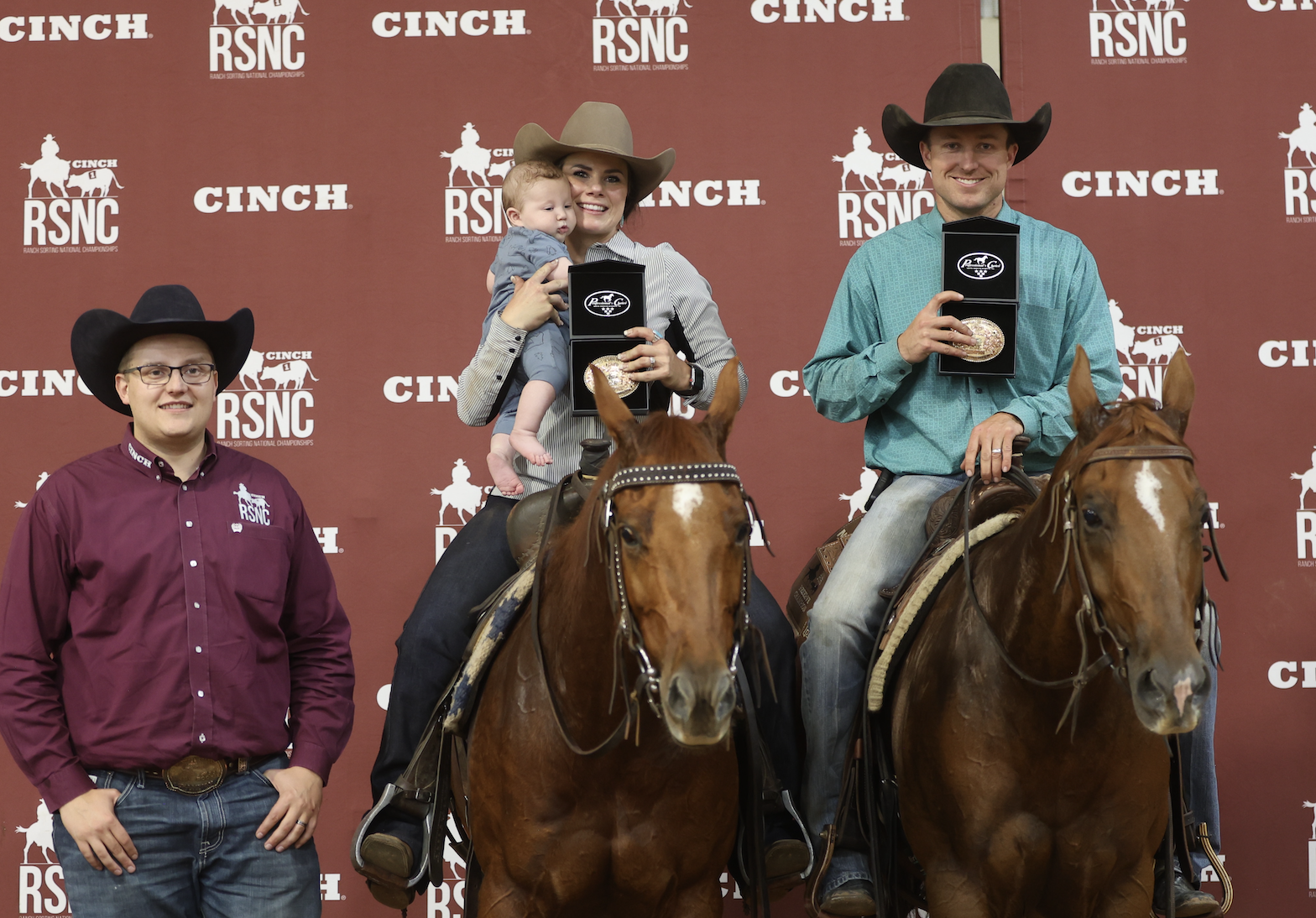 Two mounted riders hold up championship buckles, one cradling a baby, beside a man standing at a Cinch RSNC backdrop