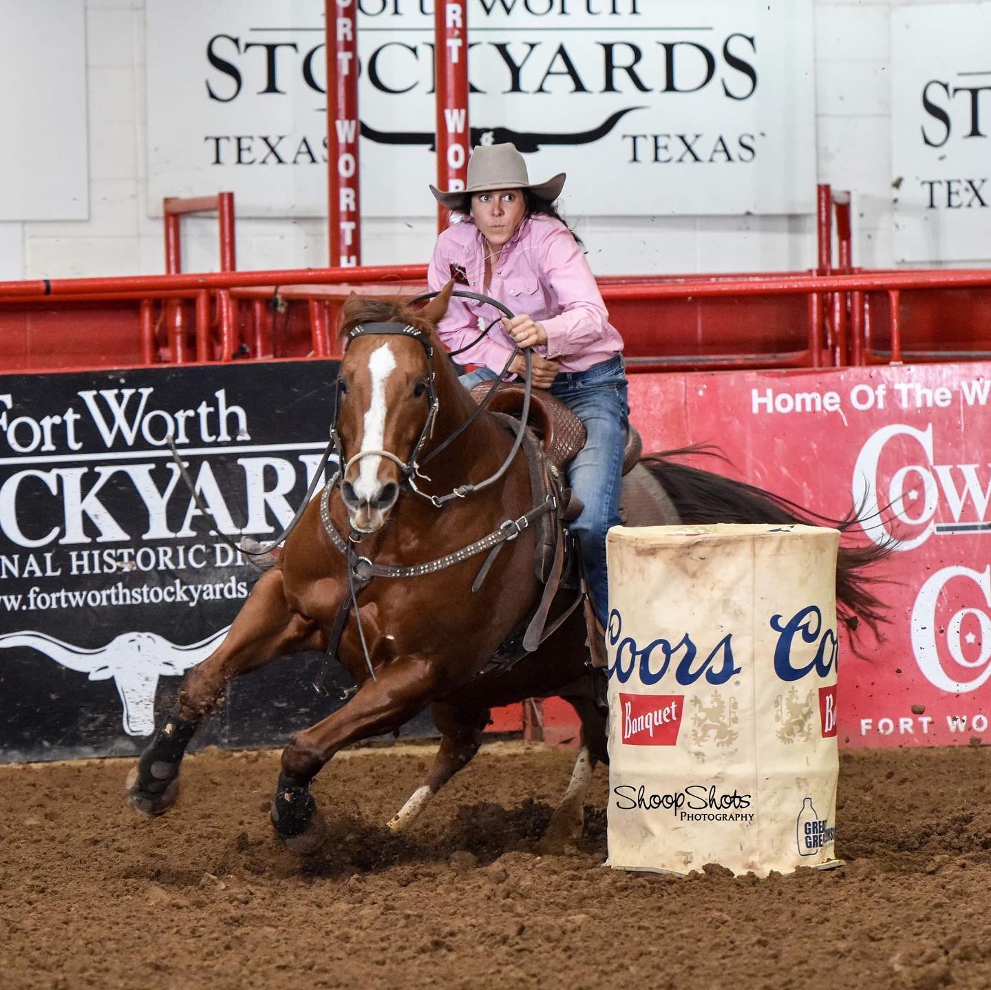 Barrel racer in a pink shirt and cowboy hat rounds a Coors barrel on a sorrel horse at the Fort Worth Stockyards