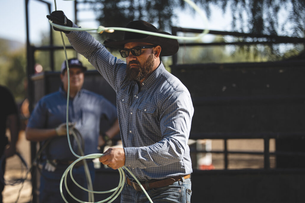 Bearded roper in a black cowboy hat and plaid shirt swings a loop overhead while holding coils of rope at an arena