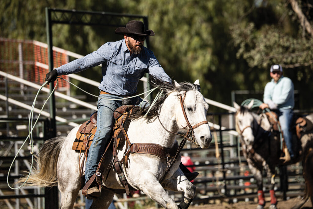 Cowboy in a black hat and blue shirt swings his rope while riding a gray horse at speed, with another rider waiting behind