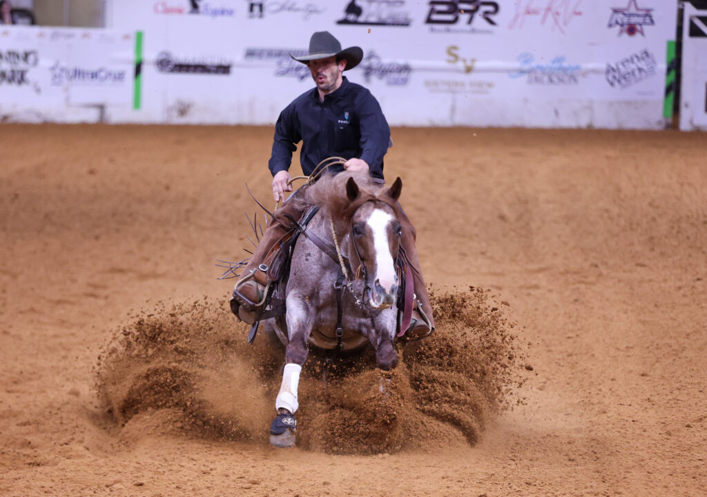 Rider in black cowboy hat slides a roan horse to a dirt-spraying sliding stop in an indoor arena