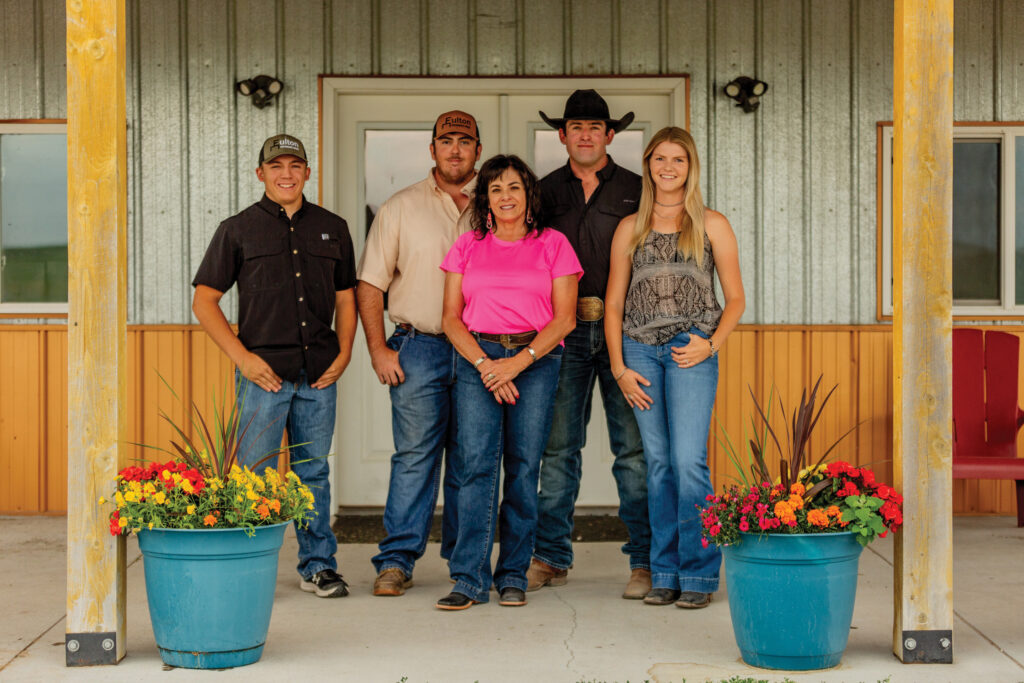 Five people in Western attire stand on a ranch porch between turquoise flower planters, smiling at the camera