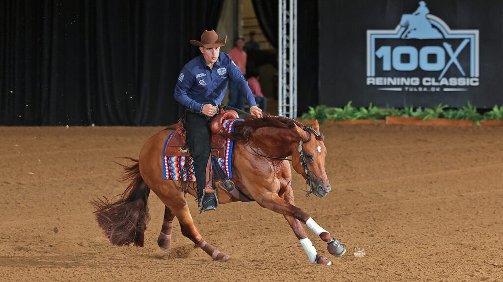 Reiner in a blue shirt and cowboy hat slides his sorrel horse to a stop in the arena at the 100X Reining Classic in Tulsa