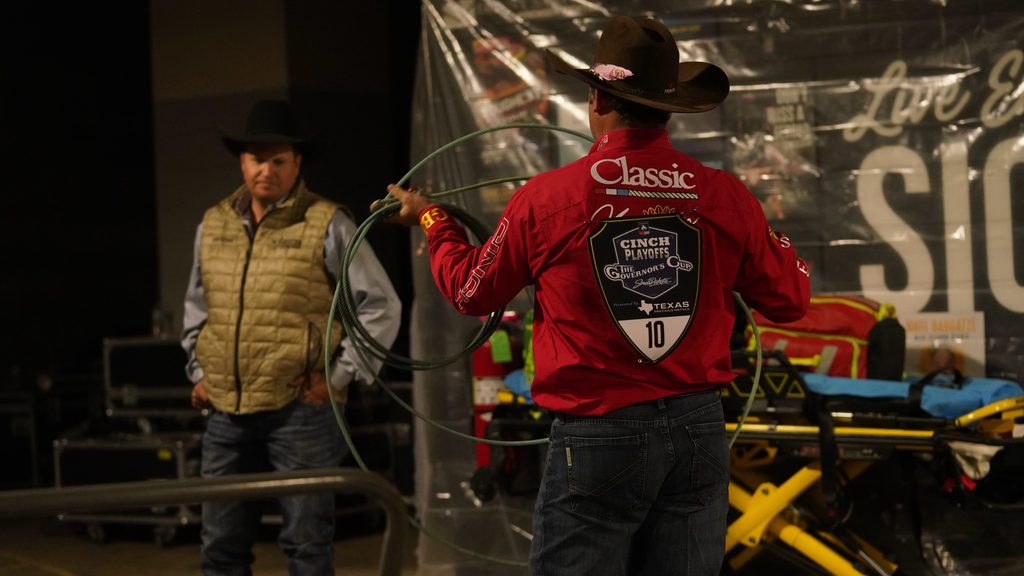 Cowboy in red jacket with number 10 practices roping while another competitor watches at an indoor rodeo event