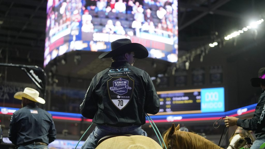 Cowboy on horseback wearing a black jacket with a Cinch Playoffs patch sits in an arena during a ranch sorting competition, with a large video screen and crowd visible in the background