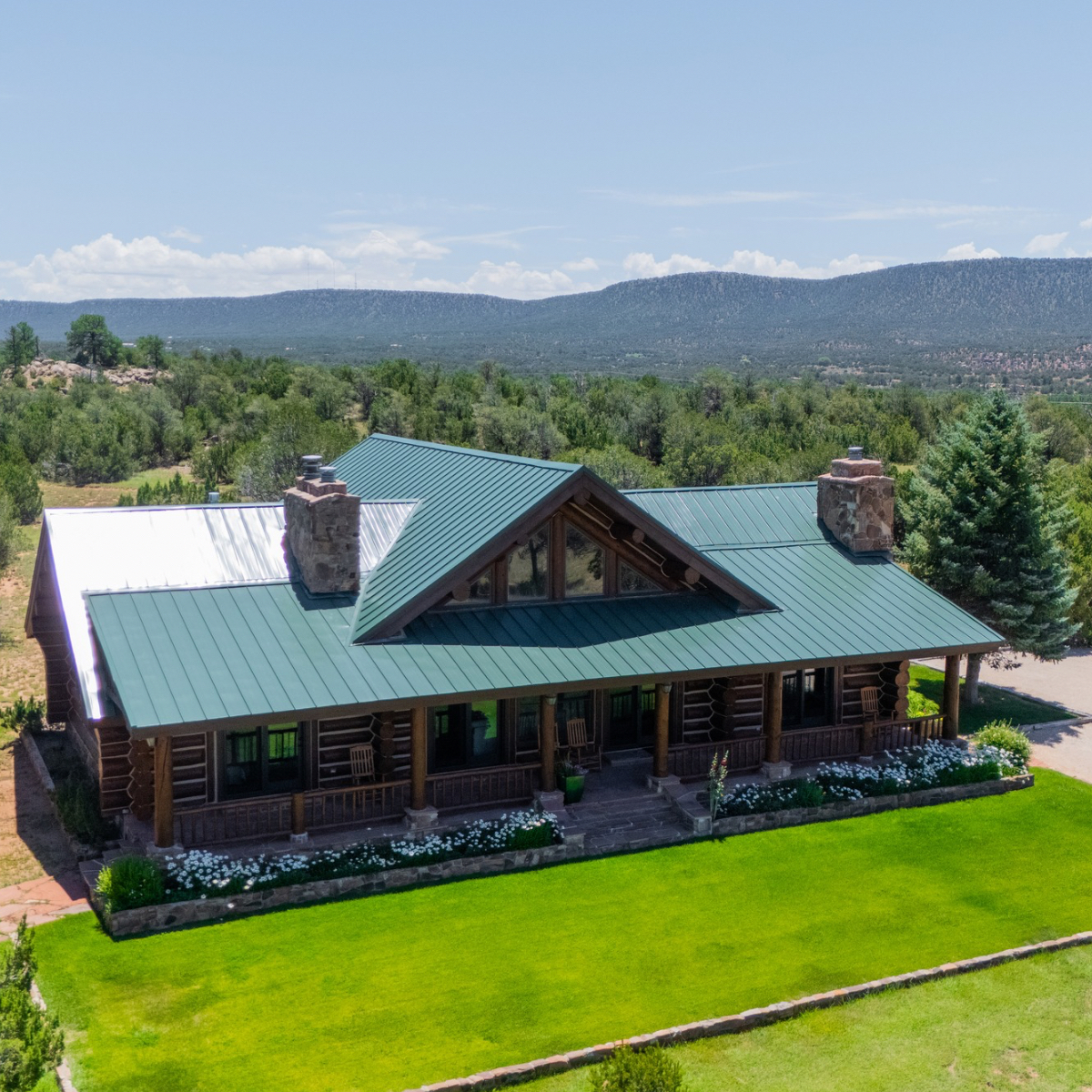 Modern log cabin with turquoise metal roof and stone chimneys sits on manicured grounds overlooking forested mountains