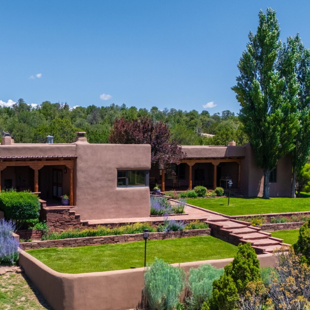 Southwestern-style adobe ranch compound with manicured grounds, featuring connected terracotta buildings with wooden portal entries and landscaped gardens under clear blue sky